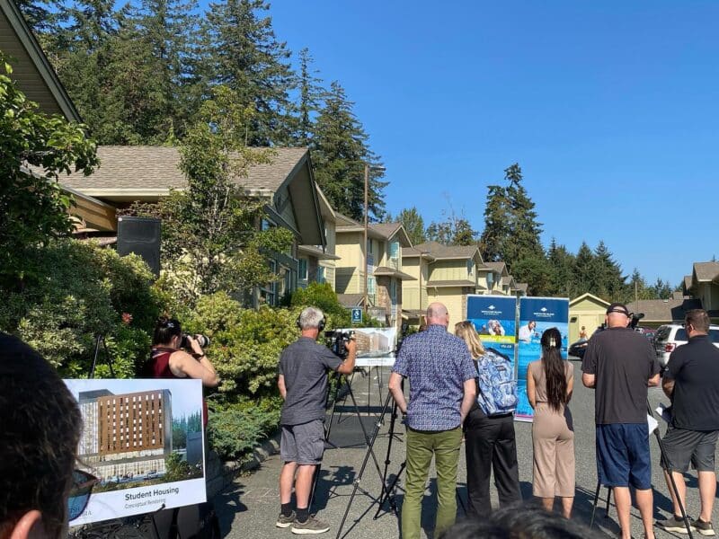 A gathering of people stand in a parking lot in front of dorm buildings as they listen to an announcement.