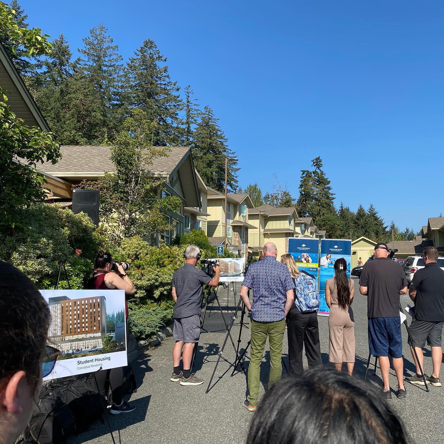 A gathering of people stand in a parking lot in front of dorm buildings as they listen to an announcement.
