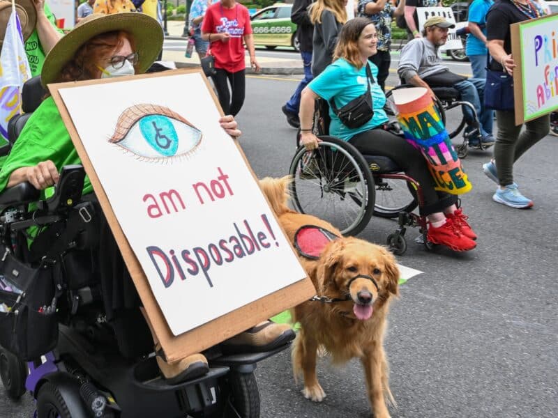 Disabled advocates hold up signs reading "we are not disposable." Two are in wheelchairs, one with crutches and others walk unassisted. One wheelchair user has a service dog.