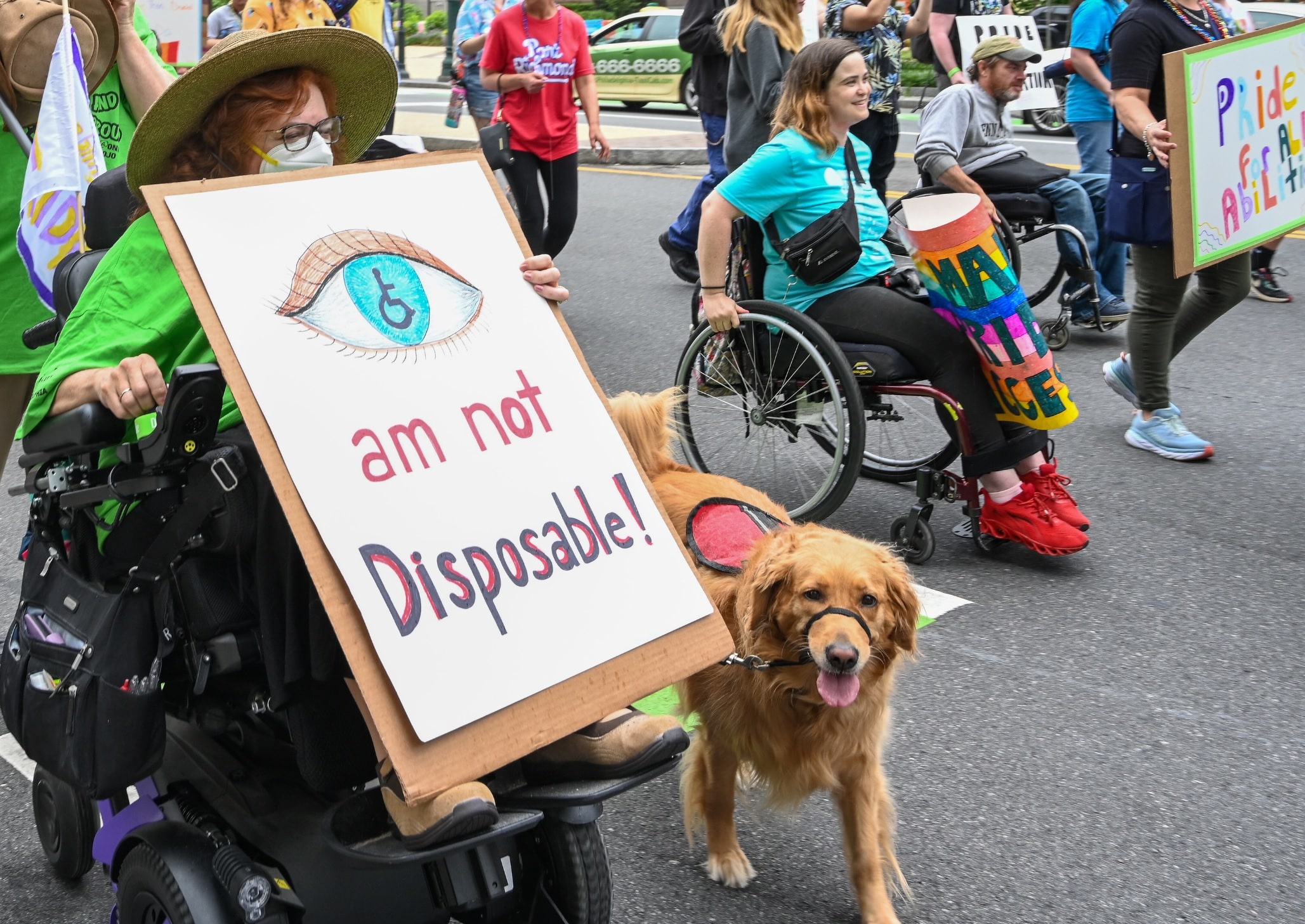Disabled advocates hold up signs reading "we are not disposable." Two are in wheelchairs, one with crutches and others walk unassisted. One wheelchair user has a service dog.
