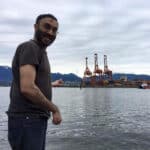Gurpreet Kambo stands in the foreground of the photo smiling at the camera, with his body turned to the side and pointing at barges and ocean behind him.