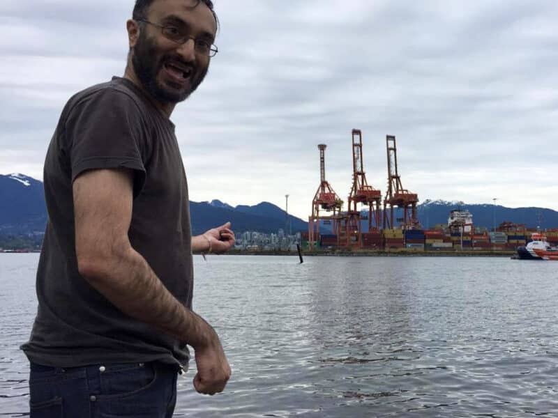 Gurpreet Kambo stands in the foreground of the photo smiling at the camera, with his body turned to the side and pointing at barges and ocean behind him.