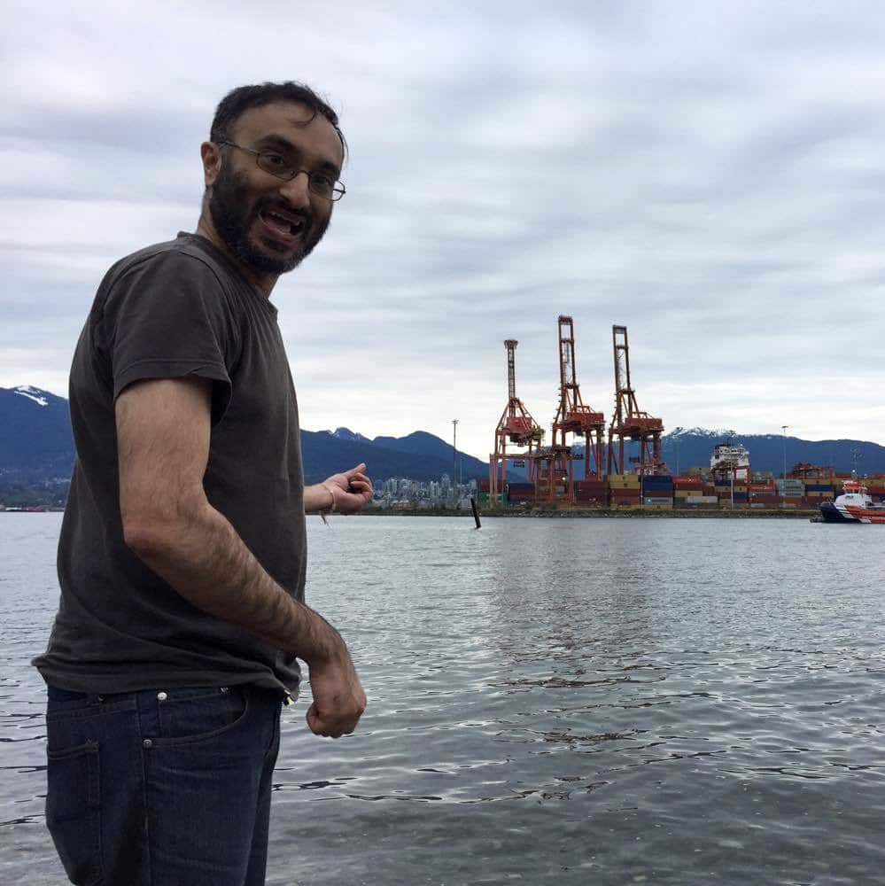 Gurpreet Kambo stands in the foreground of the photo smiling at the camera, with his body turned to the side and pointing at barges and ocean behind him.