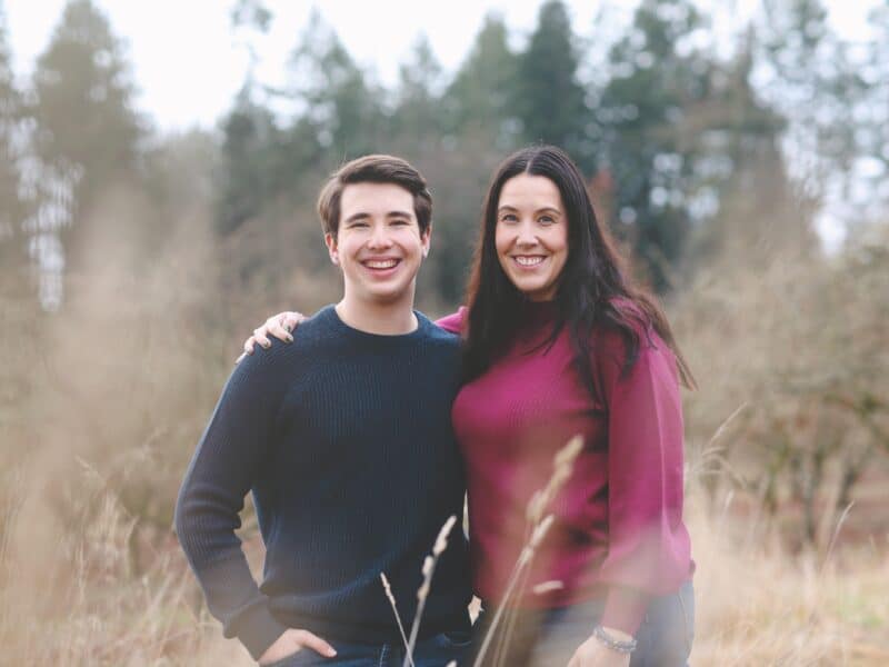 A photo of Ryland Racicot (left) and his mother Sylvia Webb (right) standing side-by-side with their arms around each other. They're smiling and facing the camera and appear to be in a field with tall grass and trees in the background.