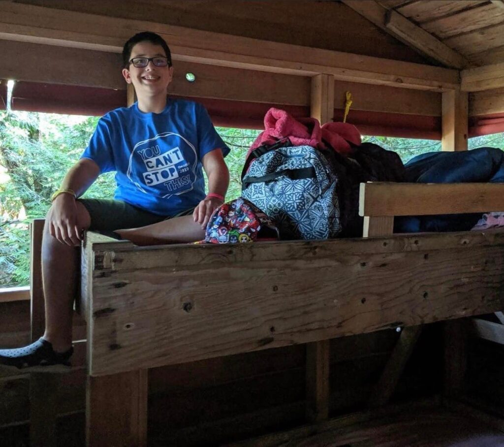 A past photo of Ryland Racicot as a camper at Camp Qwanoes. He wears a Camp Qwanoes t-shirt and sits on the top bunk of a wooden bunk bed in a cabin, smiling.