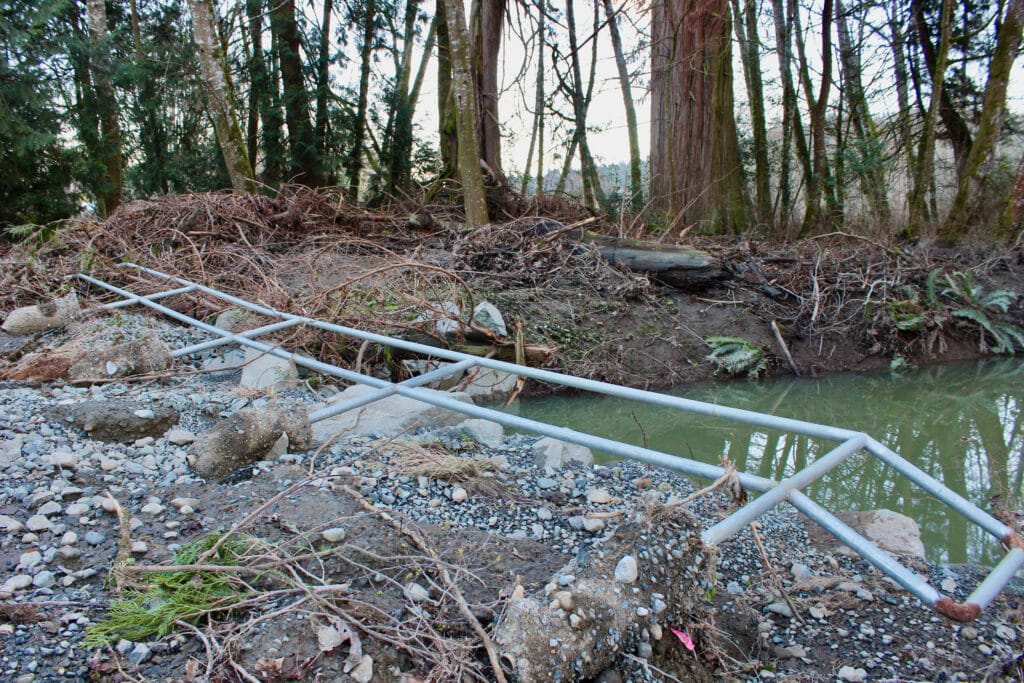 A metal barrier and forest debris is seen scattered along a river bank.