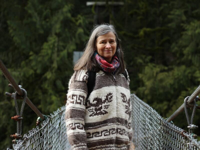 A photo of Heather Pritchard from waist-up, standing on a suspension bridge and smiling towards the camera. Dark green forest is seen behind her.