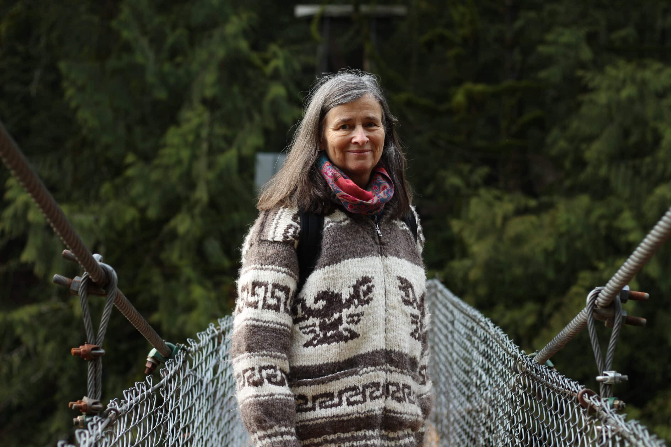 A photo of Heather Pritchard from waist-up, standing on a suspension bridge and smiling towards the camera. Dark green forest is seen behind her.