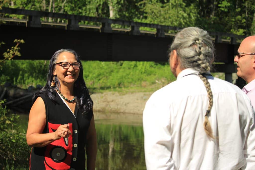 Chief Lydia Hwitsum stands on the left of the photo, facing two people whose backs are to the camera standing on the right side of the photo. The Xwulqw'selu Sta'lo' (Koksilah River) is behind Hwitsum.