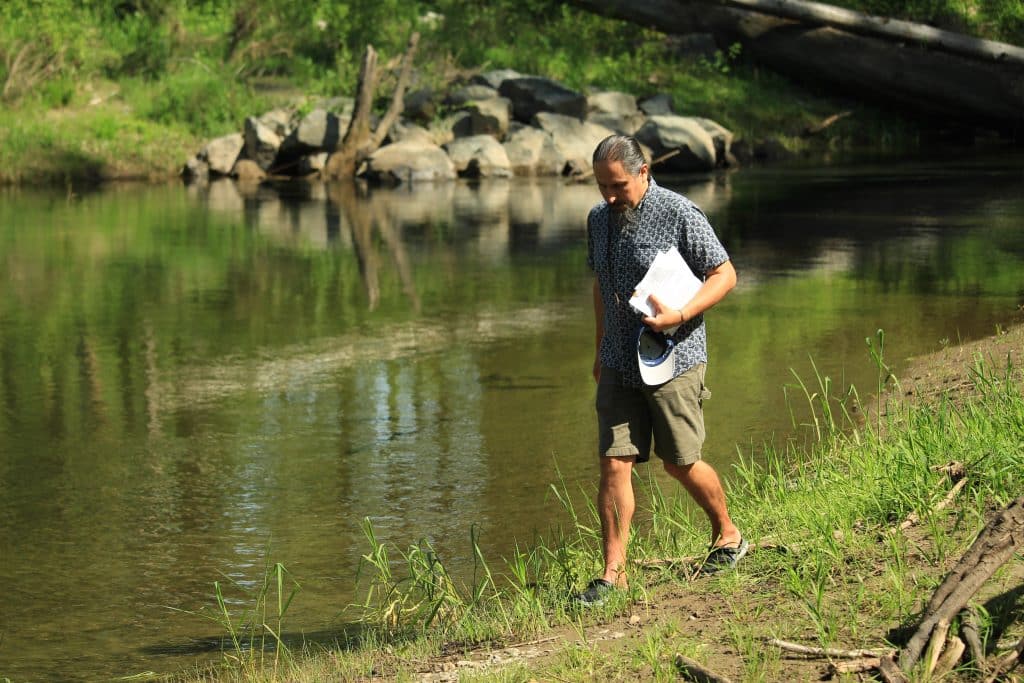 Q’utxulenuhw (Tim Kulchyski) walks along the grassy bank of the Xwulqw'selu Sta'lo' (Koksilah River). He is looking down at the ground and holds a stack of papers in his left hand.