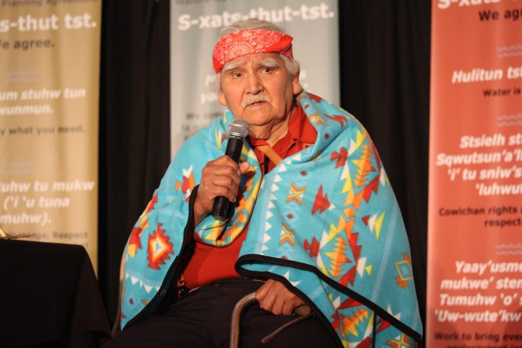 Elder Luschiim sits in a chair holding a microphone as he addresses the room. He wears a ceremonial blanket over his shoulders.