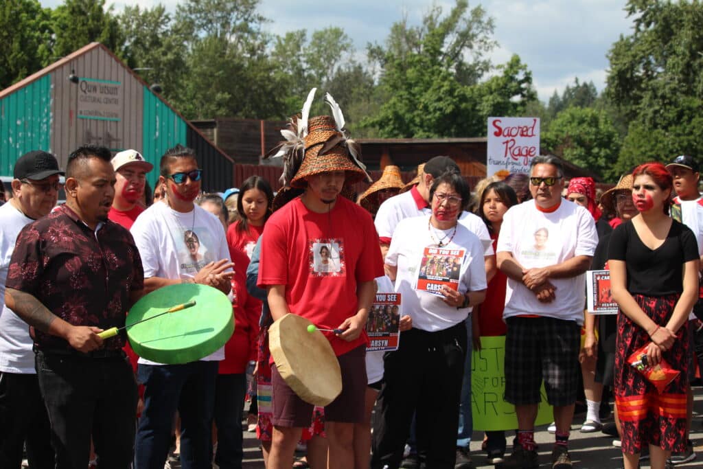 Benny George, Carsyn Mackenzie Seaweed's father, leads a march. He is drumming and wears a cedar hat and red shirt with his daughter's face on it. A crowd of community and family walk beside and behind him.