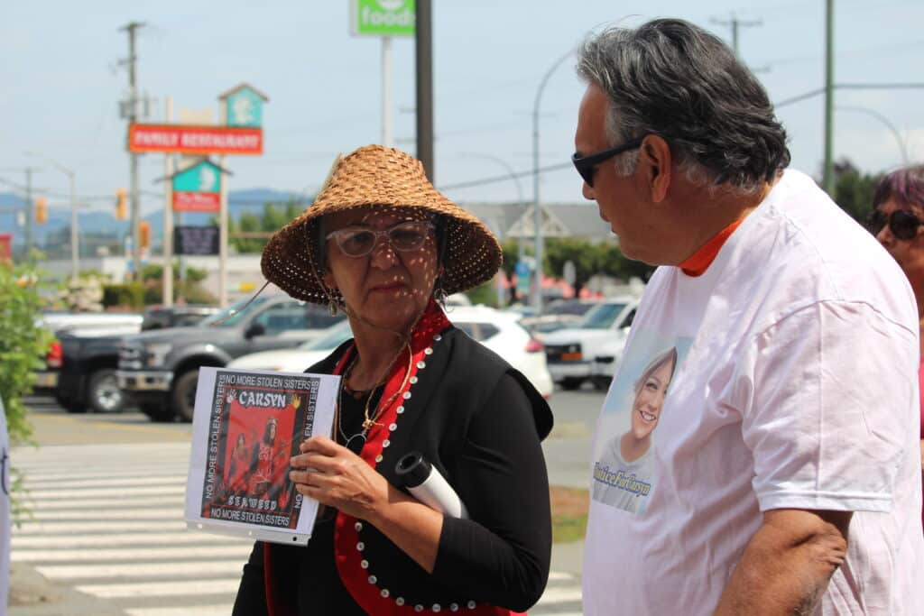 To the left of the photo is Chief Lydia Hwitsum, holding a poster that says "Carsyn Seaweed" and "No more stolen sisters." She is looking to and speaking with Chief Rick Johnson, on the right of the photo.
