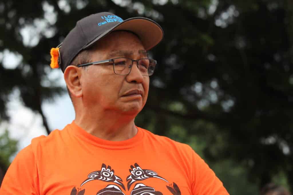 A photo of Eddie Charlie, from shoulders-up. He wears an orange shirt and black ball cap with an orange shirt pin on it.