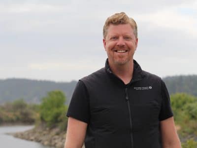 Steven Henstra is pictured, from chest up, smiling towards the camera. The Cowichan estuary and a dike along the estuary is behind him.