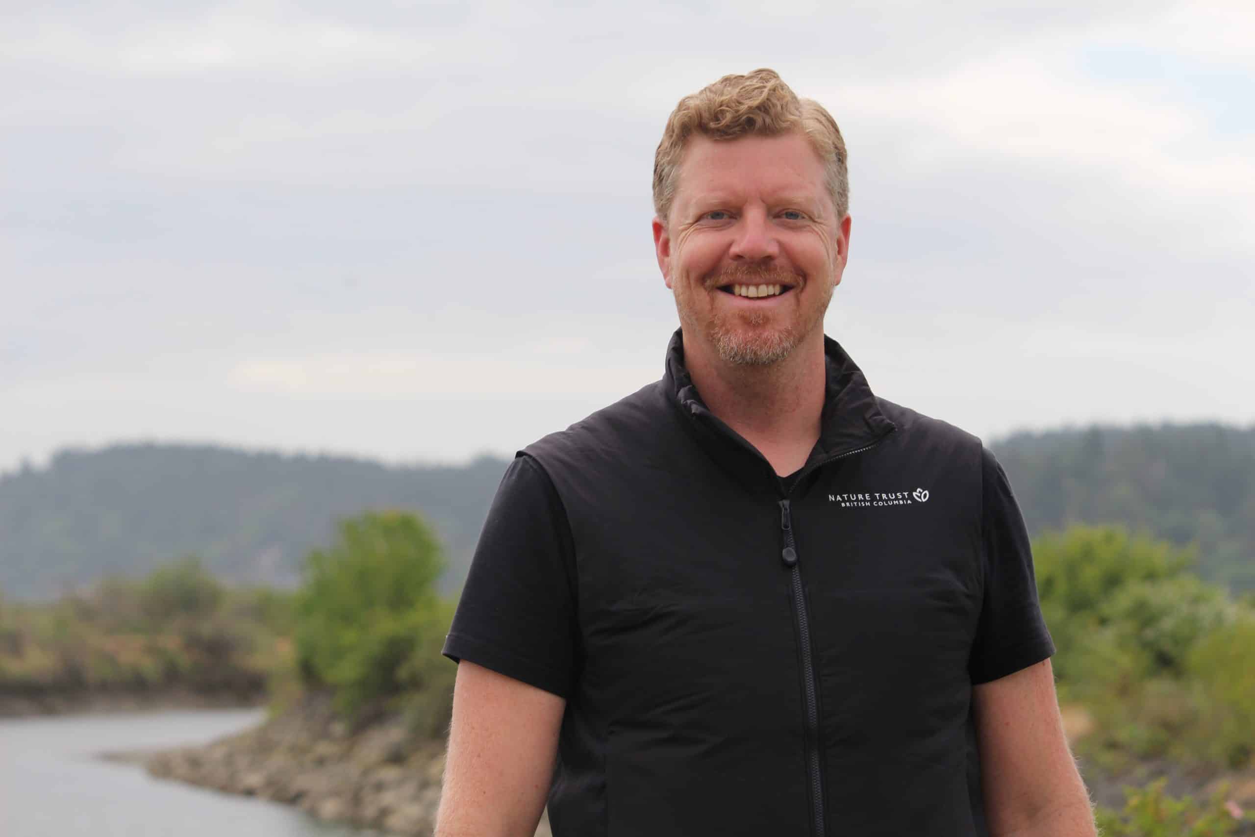 Steven Henstra is pictured, from chest up, smiling towards the camera. The Cowichan estuary and a dike along the estuary is behind him.