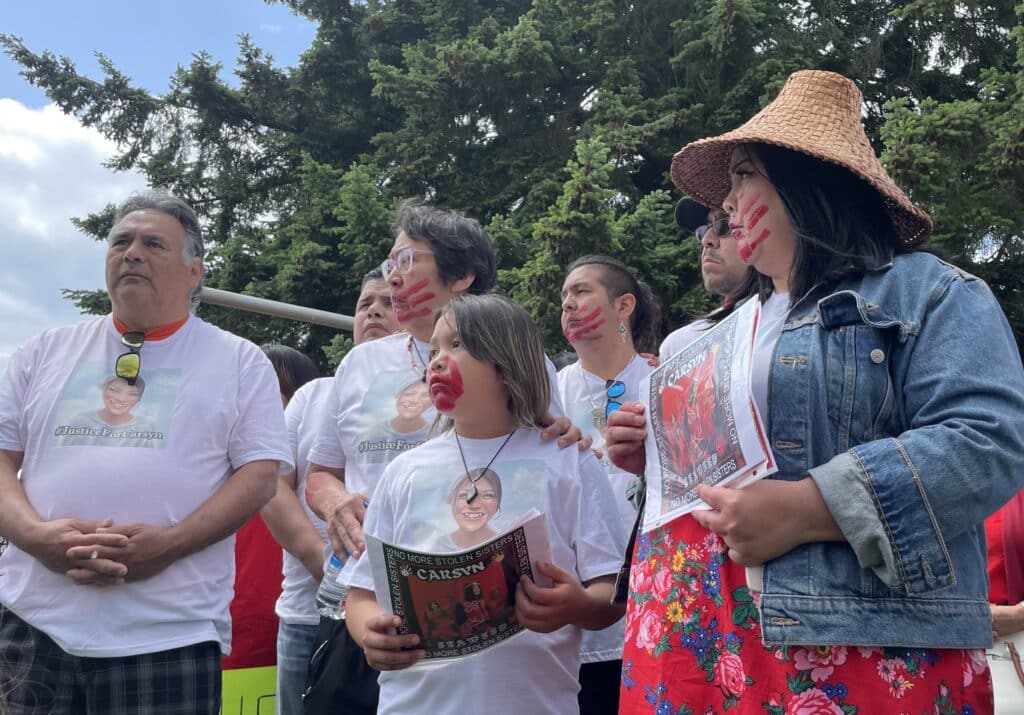 Chief Rick Johnson and Carsyn's grandma, younger sister and mother stand together, backed by family. They all wear white t-shirts with Carsyn's photo on them and the mother, grandmother and sister have red handprints painted on their faces.