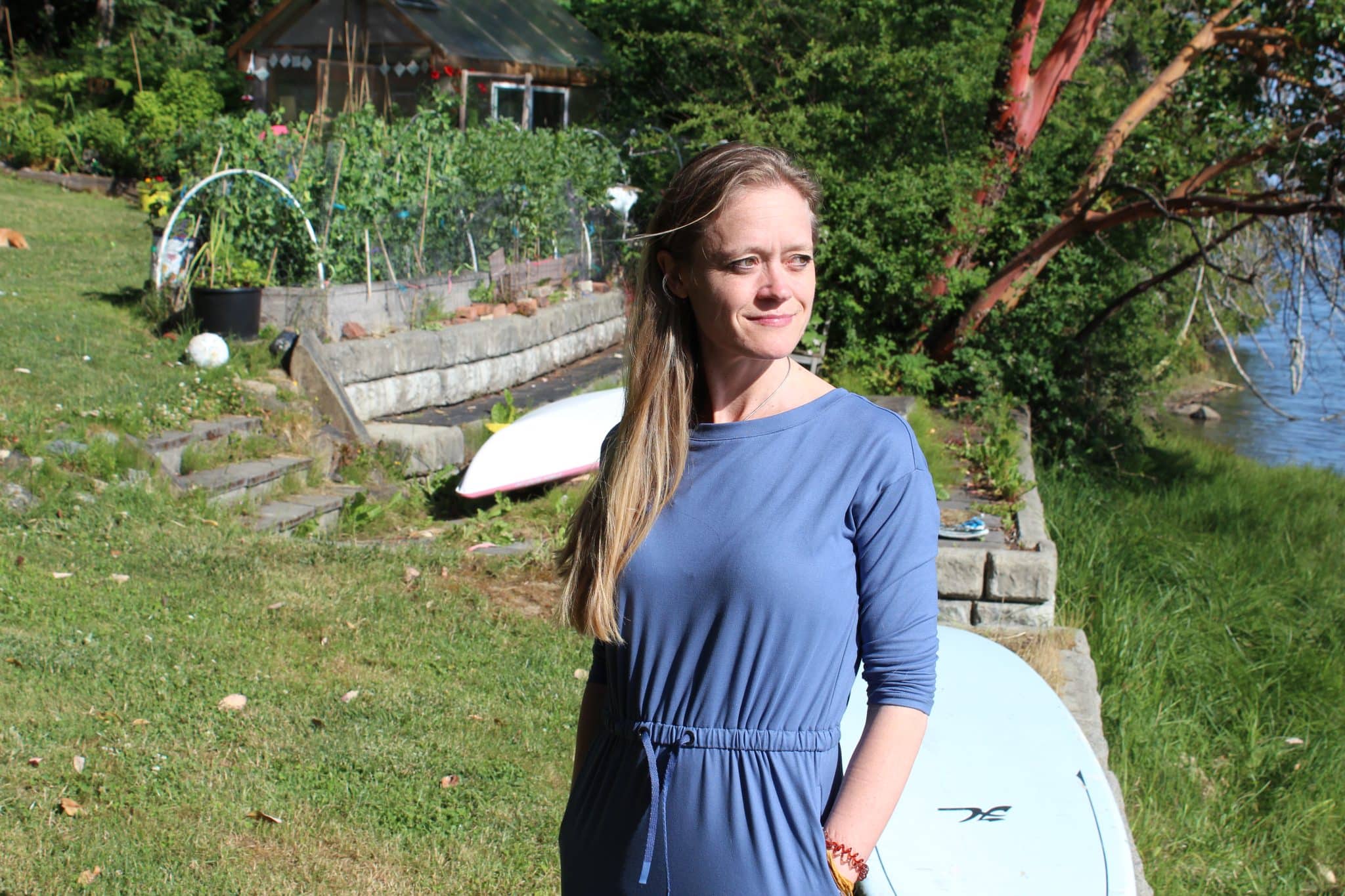 A photo of Shannon Dames. She is standing outdoors, with a garden behind her, and looking into the distance.