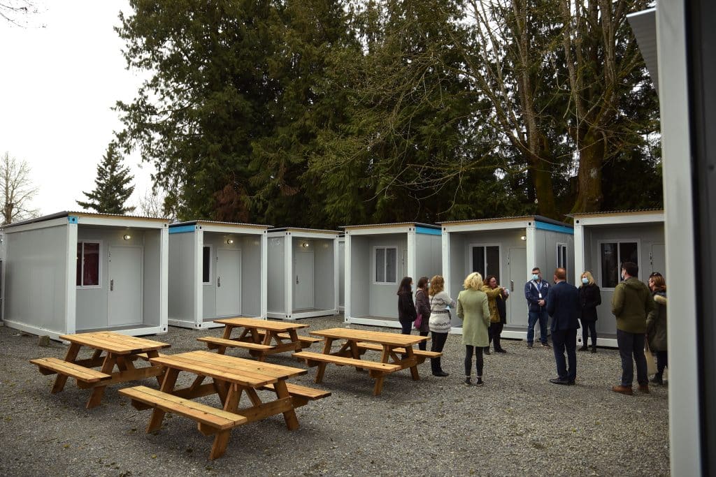 Small modular homes that look like shipping containers form a square, with their entrances facing inwards to a courtyard with four picnic tables. Off to the right of the photo is a group of people standing in a circle and speaking.