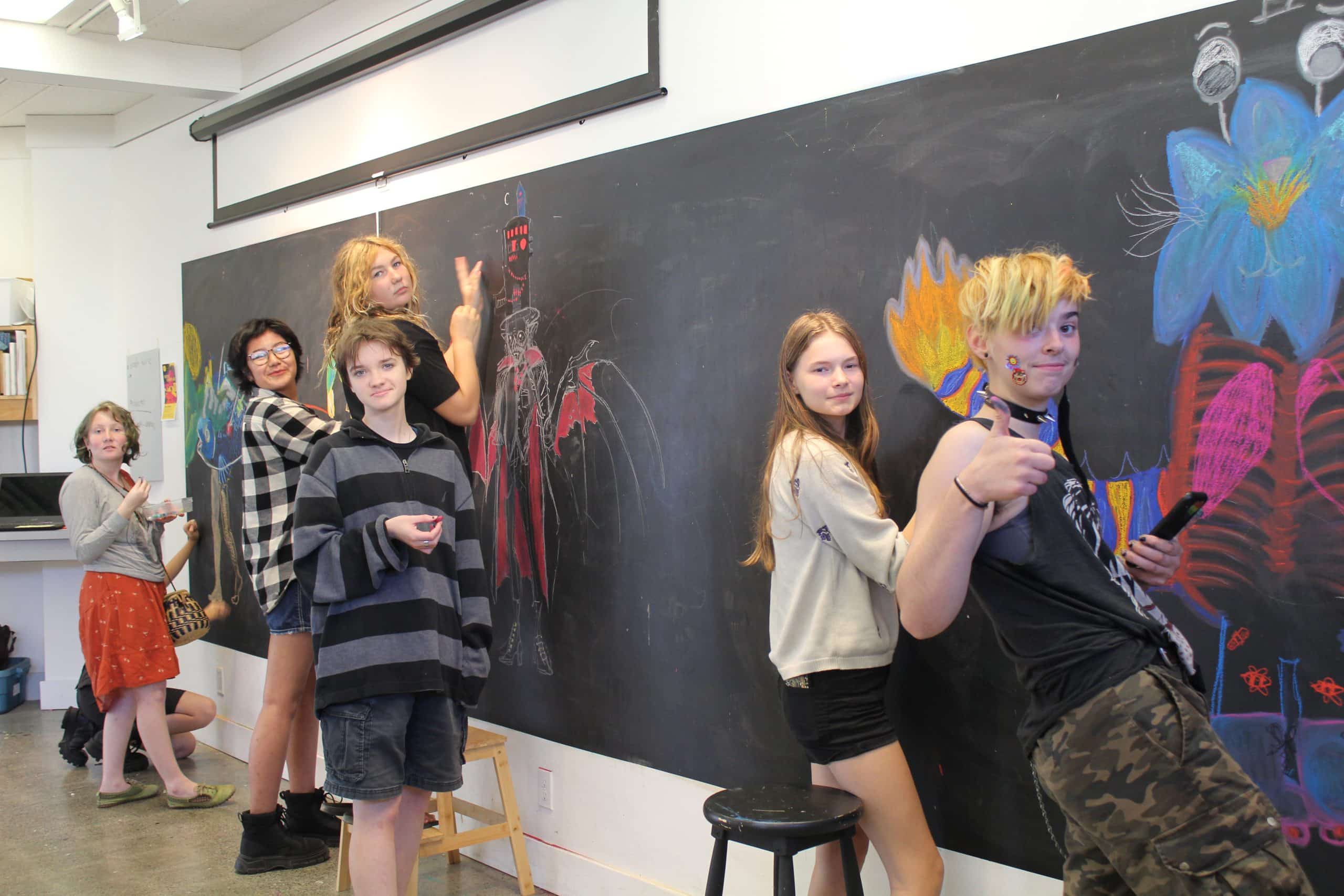 A group of young teenagers stand at a blackboard, drawing. One teen on the right gives a thumbs up.
