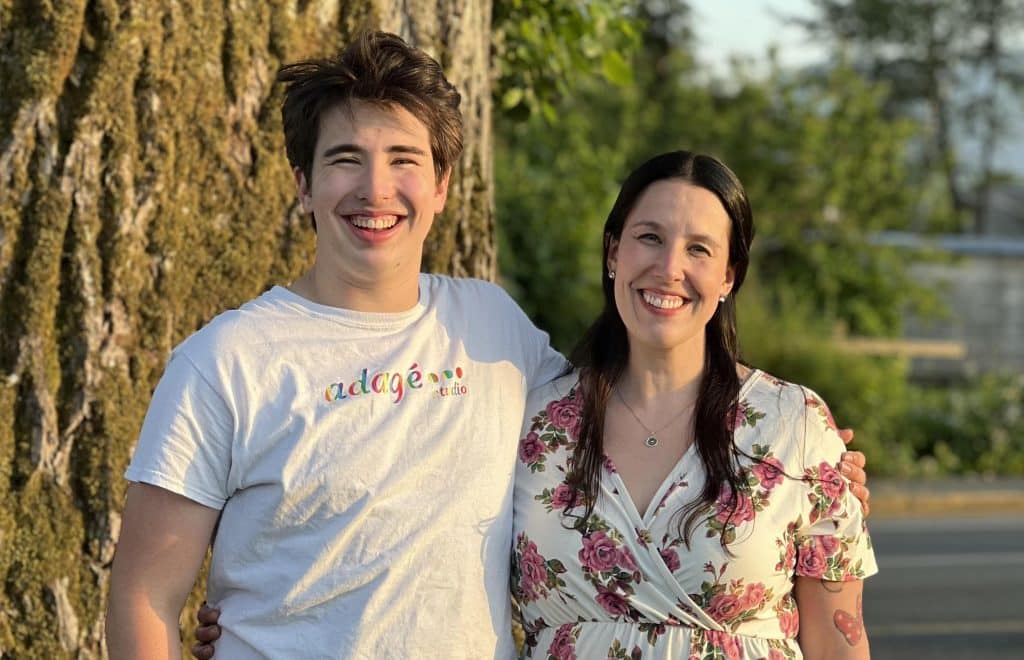A photo of Ryland Racicot, standing to the left of his mom, Sylvia Webb. Ryland's arm is around Sylvia and the photo is taken from chest-up. They are both smiling.