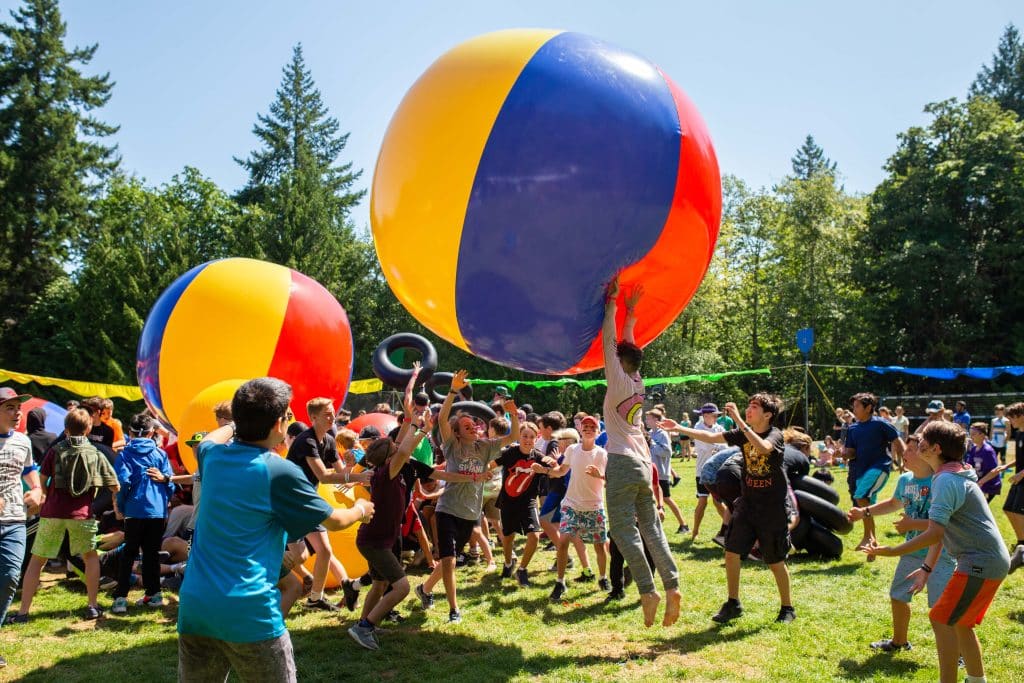 Youth are seen playing outdoors in the sun with giant, colourful beach ball-looking toys called moon balls.