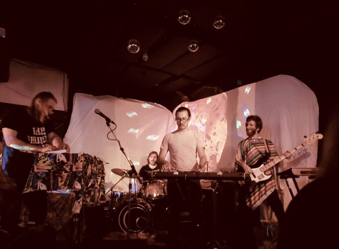A sepia tinted photo of four men playing various instruments at an indoor venue.