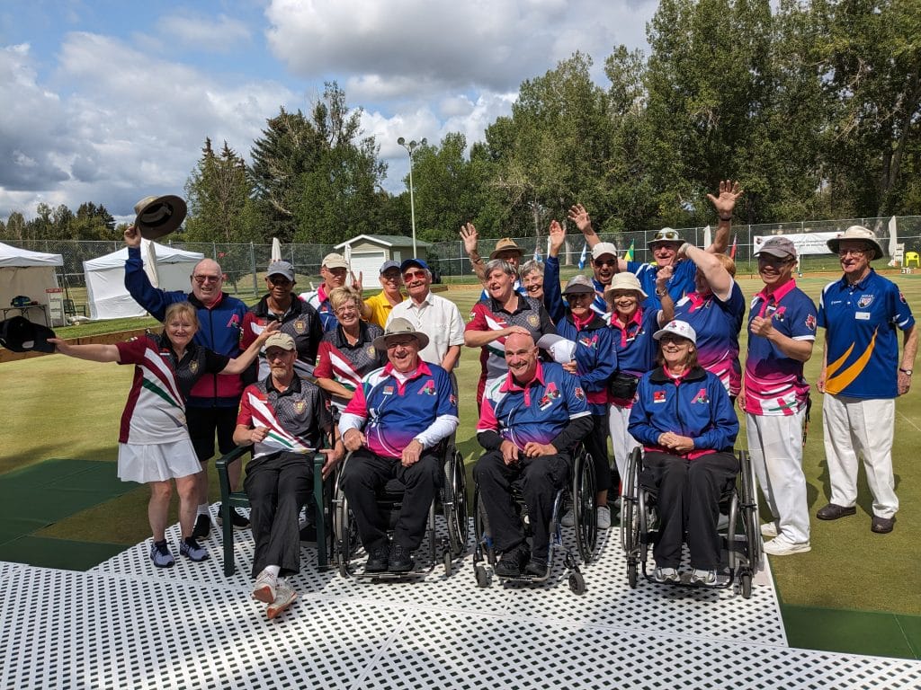 A group photo of disabled lawn bowlers at the Para-Bowl Championships in Calgary. Some are elderly, some are in wheelchairs, many are cheering and tossing hats and throwing their arms up, laughing and smiling.