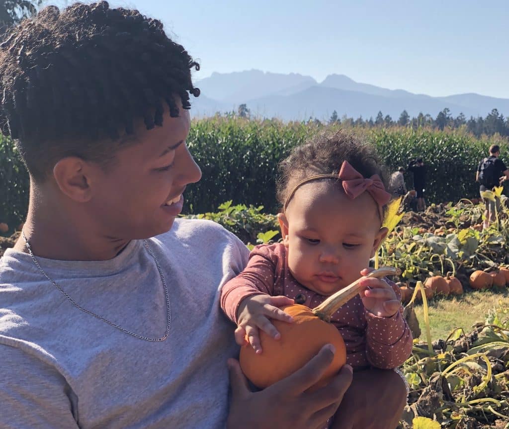 Max is shown holding his baby sister Chloe in a pumpkin patch, he's looking at her smiling as she holds the stem of a small pumpkin.
