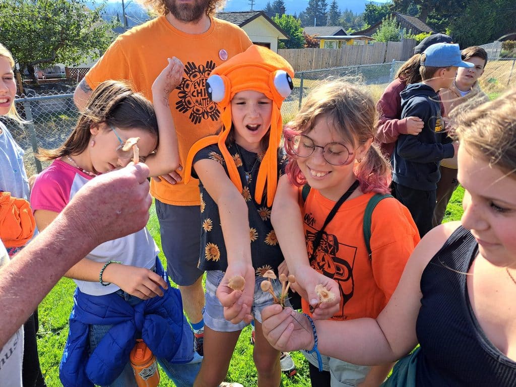 A group of smiling kids in a circle hold up mushrooms they found during recess.