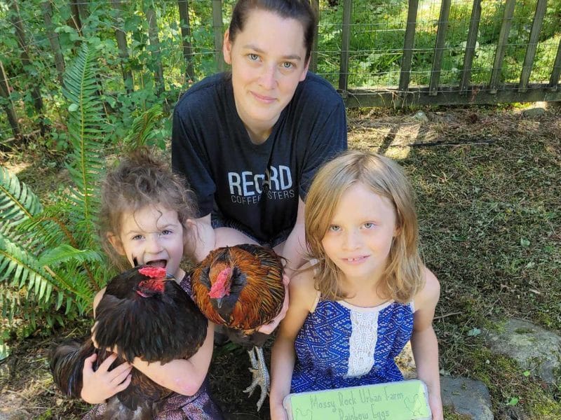 A woman with a bun in her hair squats behind her two children, one of whom is curly haired and holding a chicken, the other who is red haired and holding a dozen eggs in a box.