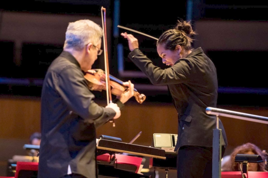 Dark-haired woman holds a conductor's baton and directs the grey-haired man she is facing, whose back is to the camera and is playing fiddle