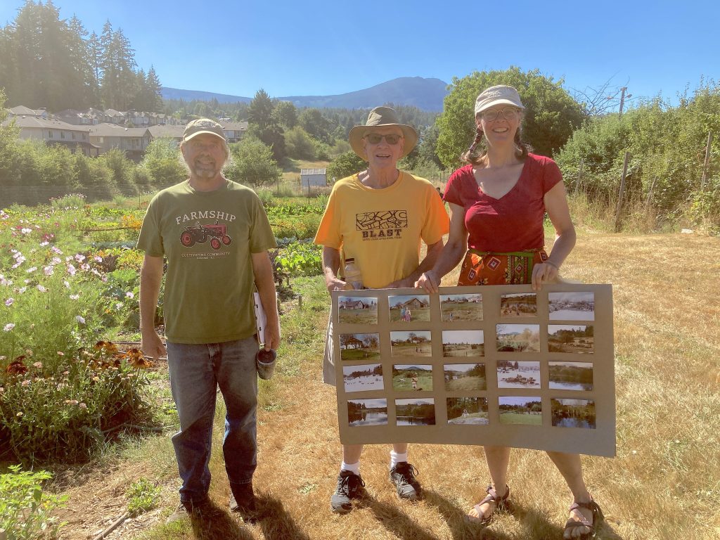 Three people stand together with hats on and a poster board of photos before a field of flowers and vegetables