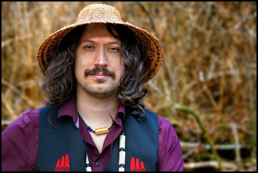 Musician Saltwater Hank looks into the camera wearing a cedar woven hat and beaded necklace
