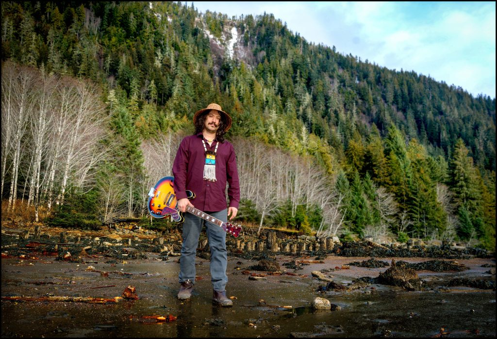 Musician Saltwater Hank stands on a beach holding a guitar and wearing a woven cedar hat