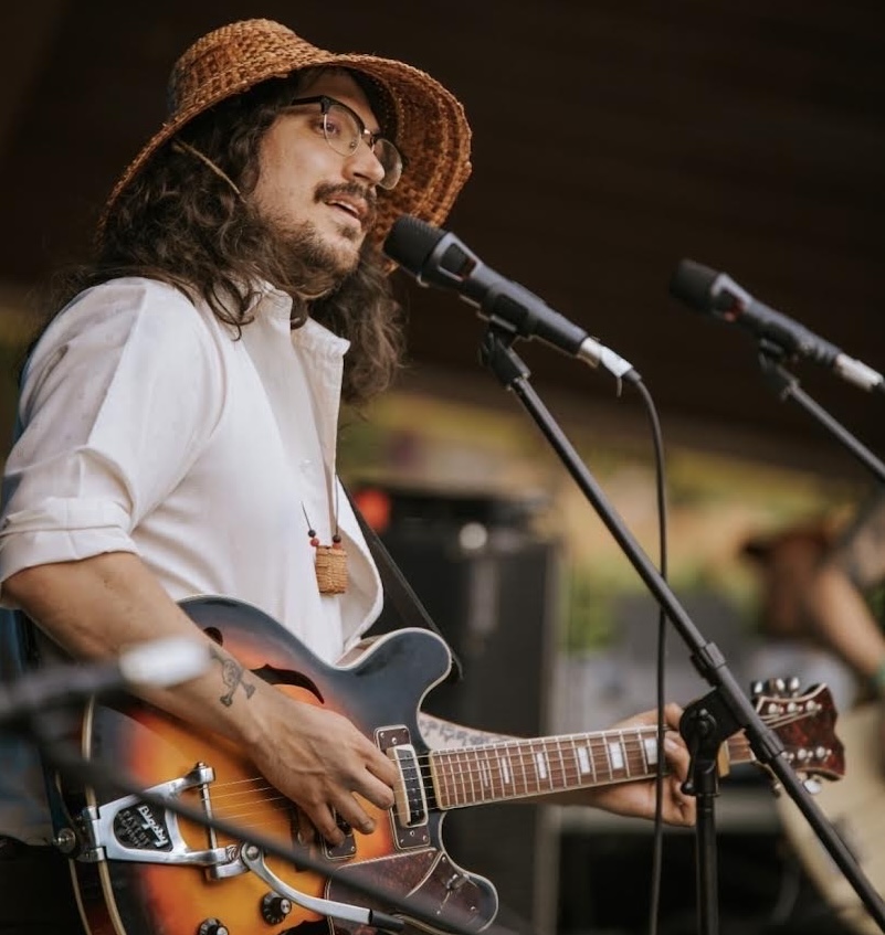 Musician Saltwater Hank sings and plays guitar wearing a cedar woven hat and white button up shirt
