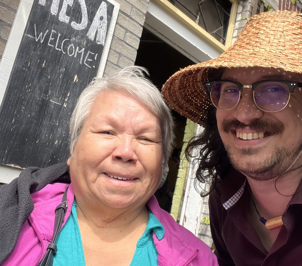 A Ts'msyen elder and musician Saltwater Hank pose together, smiling. He is in a woven cedar hat