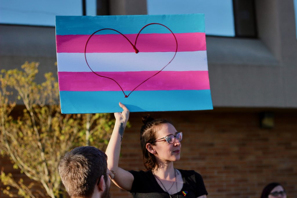 A person holds a handmade sign above their head that has been painted the colours of the trans flag. A heart is drawn on top in black.