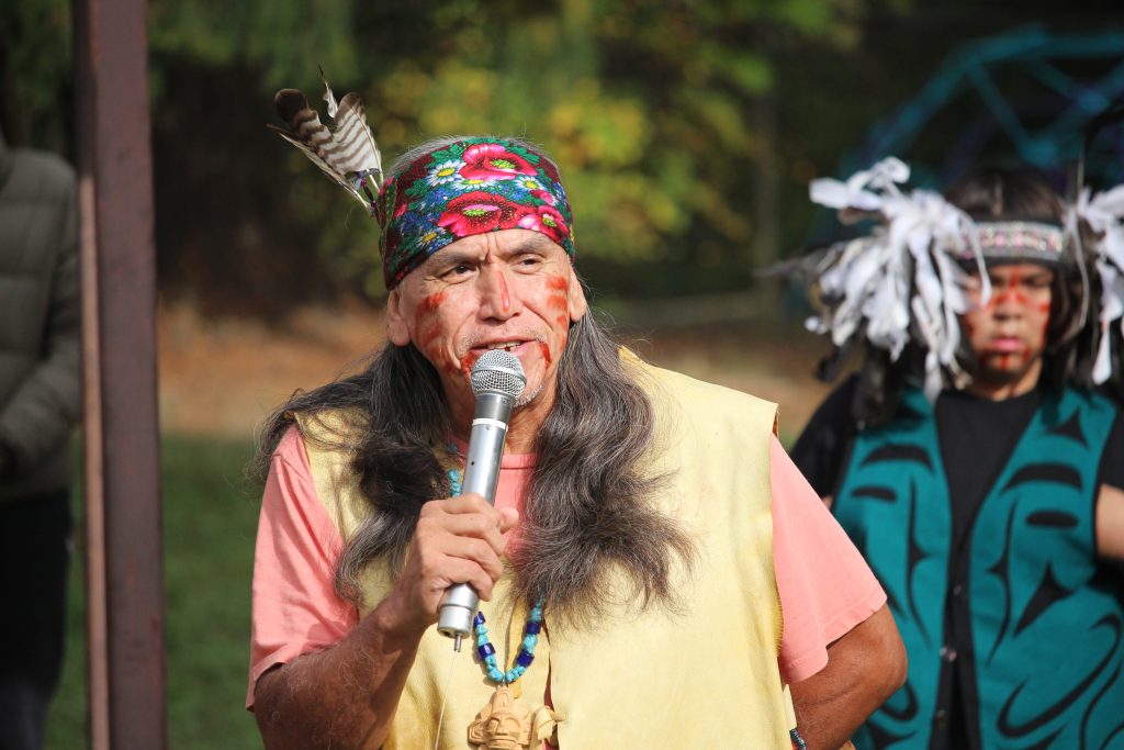 Qwiahwultuhw dressed in regalia, speaks into a microphone.