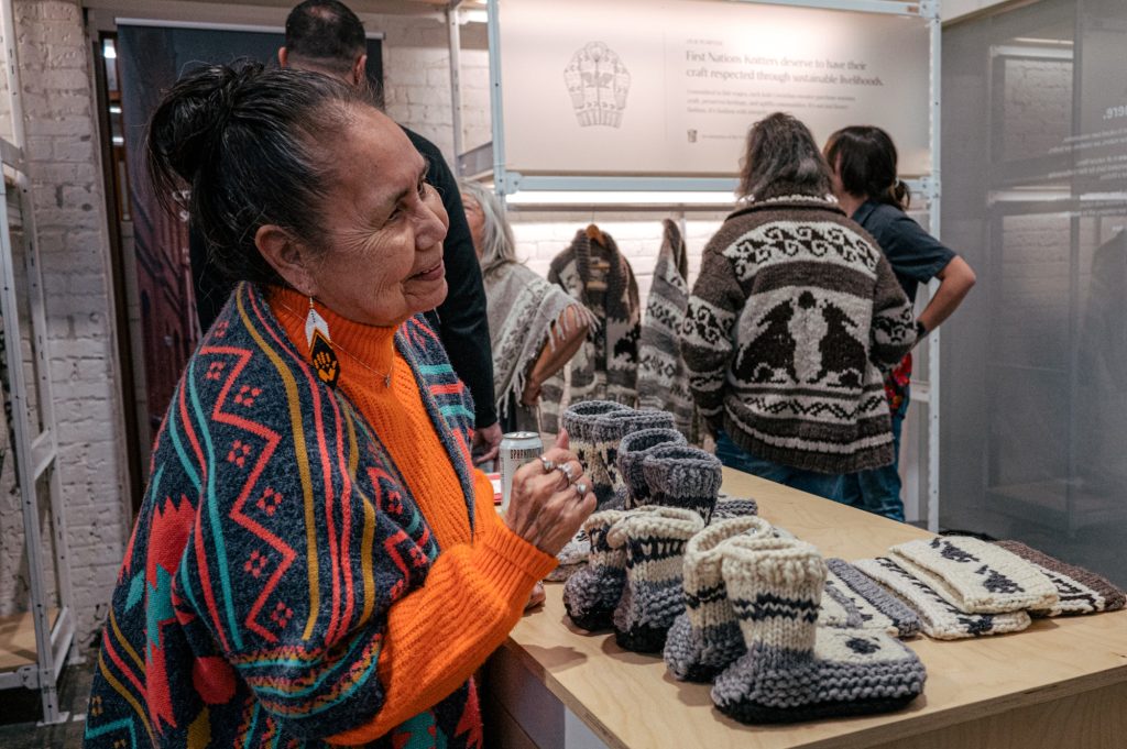 A woman smiles near a table with knitted slippers and head bands
