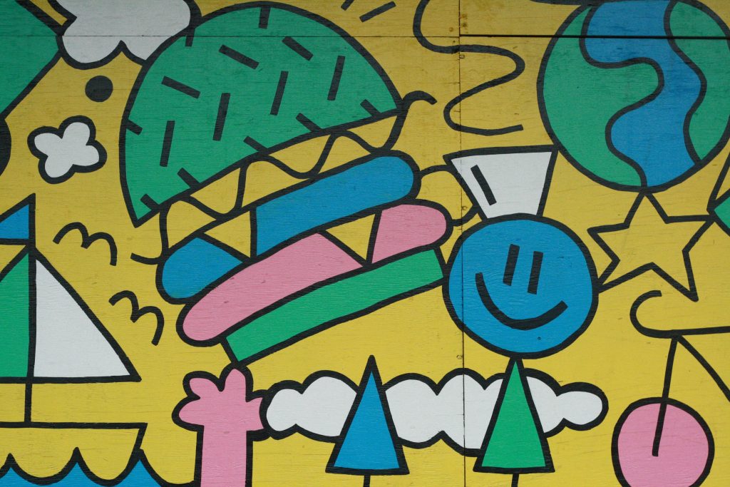 A close-up detail of a mural shows colourful green, yellow, blue and pink designs with a hamburger, clouds and a stick person with a blue smiley face and white hat.