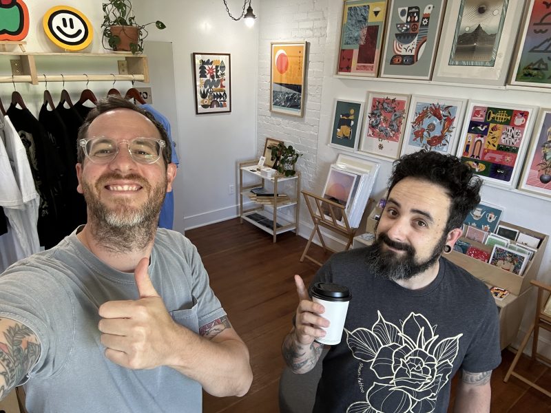 Sebastian and Mauro stand in their studio giving a thumbs up. Mauro holds a coffee cup.