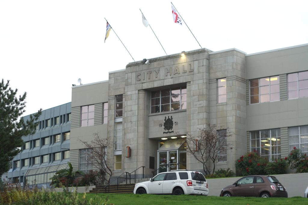 Nanaimo City Hall stands with flags under a grey sky. The building is also grey in front of a green lawn