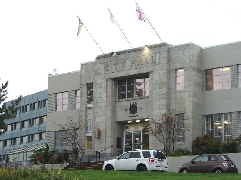 Nanaimo City Hall stands with flags under a grey sky. The building is also grey in front of a green lawn