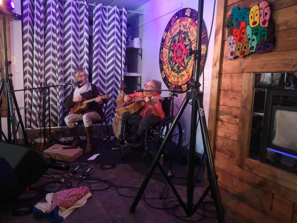 two women hold instruments and sing into microphones on a small stage