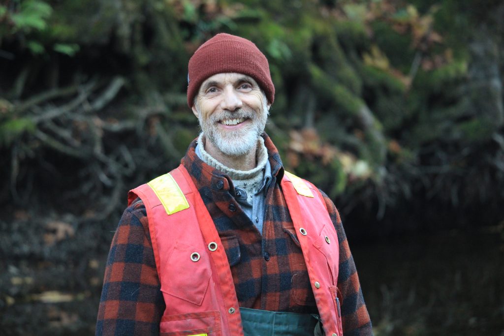 A headshot of Tom Rutherford. In the background is some forested area and ferns. He wears a brick red toque, a red and black plaid shirt, a red and yellow safety vest and the tops of his chest waders are seen as well.