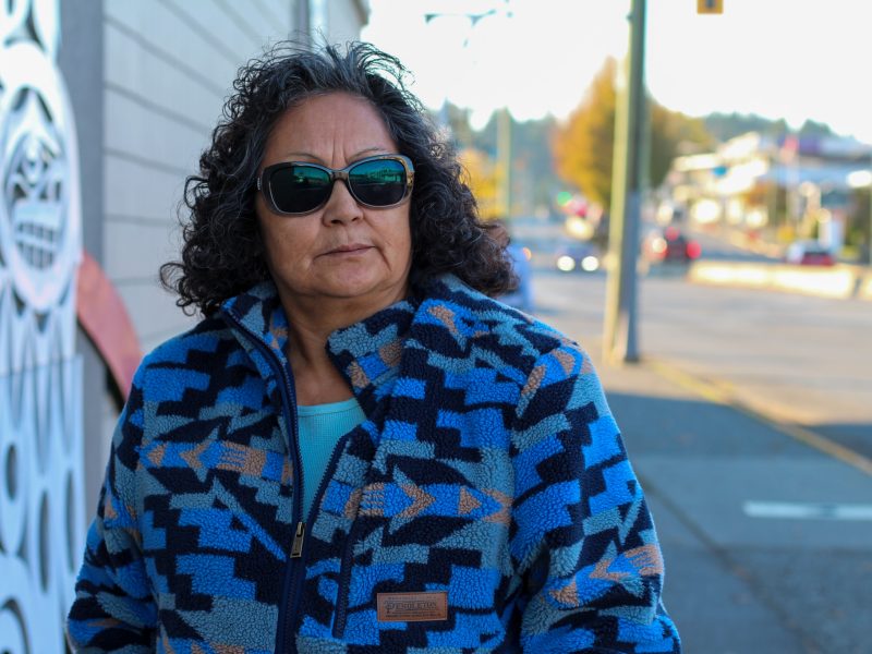 A photo of Monica Patsy Jones, taken from torso-up. She is wearing sunglasses and a zip-up sweater and is standing outdoors in Duncan.