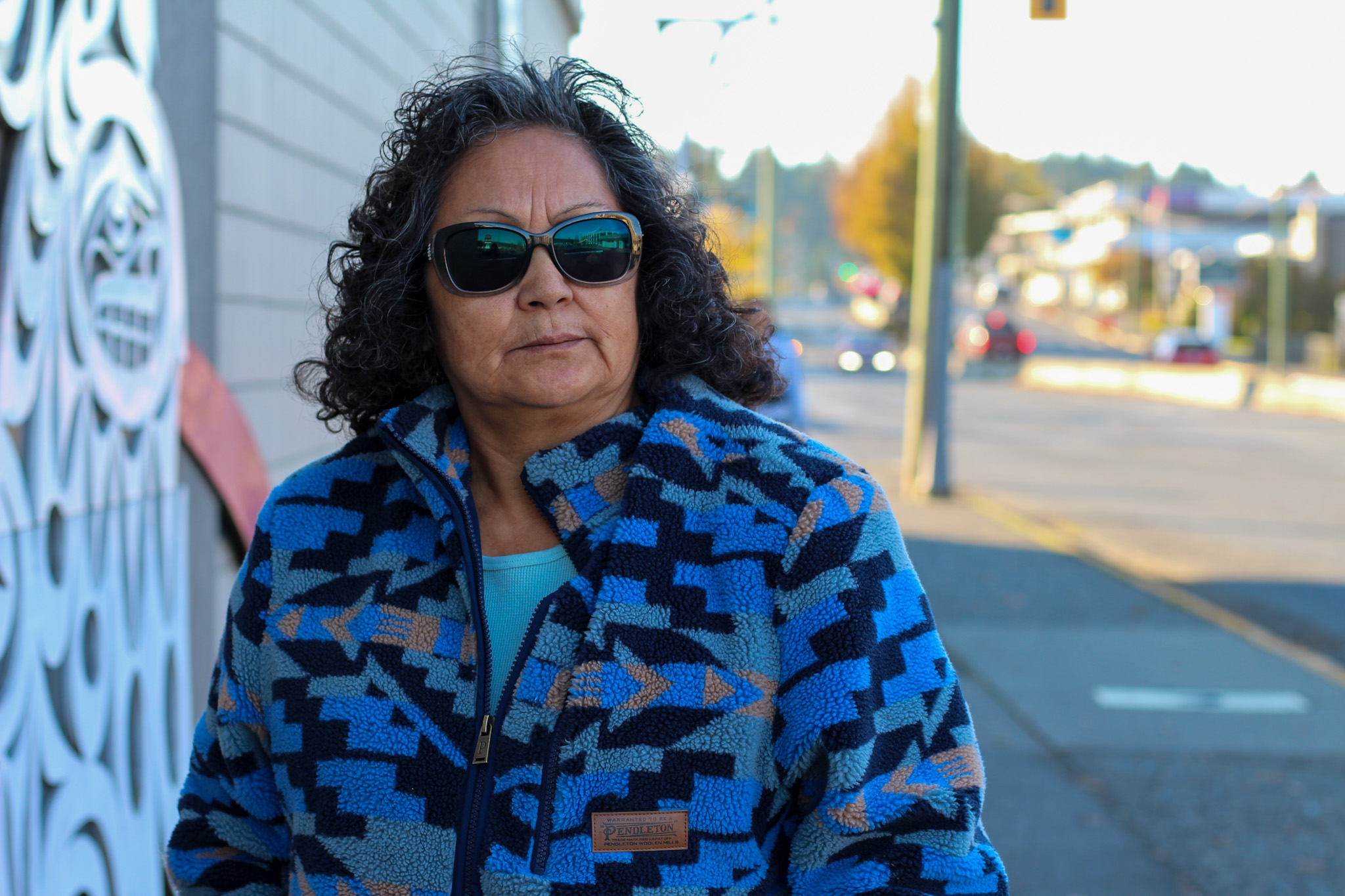 A photo of Monica Patsy Jones, taken from torso-up. She is wearing sunglasses and a zip-up sweater and is standing outdoors in Duncan.