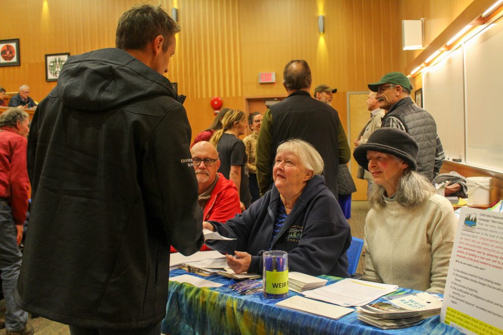 One man (left) and two women (centre and right) sit behind a table with papers and pens on it. They speak with a man (his back is to the camera) and the woman in the centre hands him a paper. Behind them is a lecture hall full of people, talking.