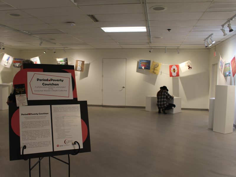 A photo of the gallery space where the Period Poverty Cowichan exhibit is. A sign on an easel at the entrance of the exhibit reads "Period Poverty Cowichan" and behind it is the exhibit, with images hung on clothesline draped along the walls. One person in the background crouches to write something on a comment sheet and two others are seen to the left in the background.